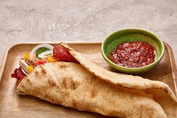 Burrito frais avec poulet et légumes à bord près de sauce chili sur fond gris béton — Photo de stock