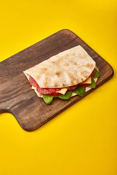 Fresh sandwich with salami, pita, vegetables and cheese on wooden cutting board on yellow background — Stock Photo