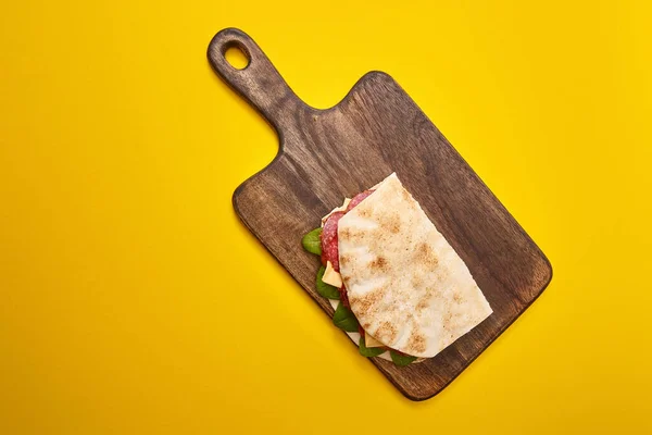 Top view of fresh sandwich with salami, pita, vegetables and cheese on wooden cutting board on yellow background — Stock Photo