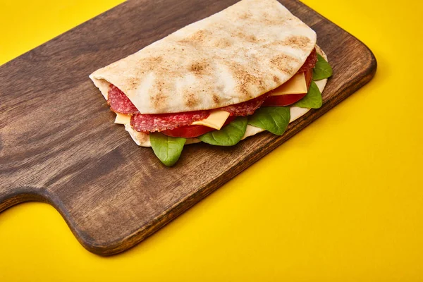 Sanduíche fresco com salame, pita, legumes e queijo em tábua de corte de madeira em fundo amarelo — Fotografia de Stock