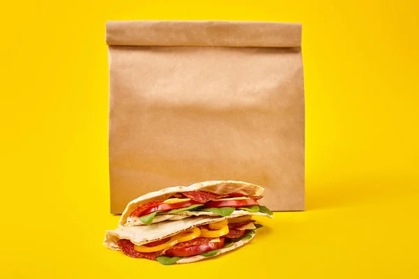 Bocadillos frescos con salami, pita, verduras y queso sobre fondo amarillo cerca de una bolsa de papel para llevar - foto de stock