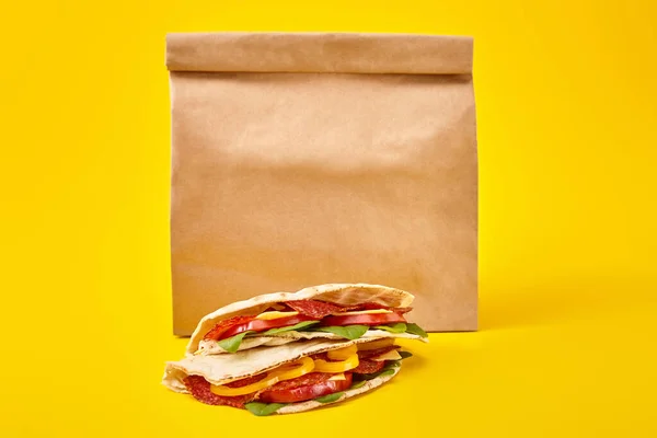 Fresh sandwiches with salami, pita, vegetables and cheese on yellow background near paper takeaway bag — Stock Photo