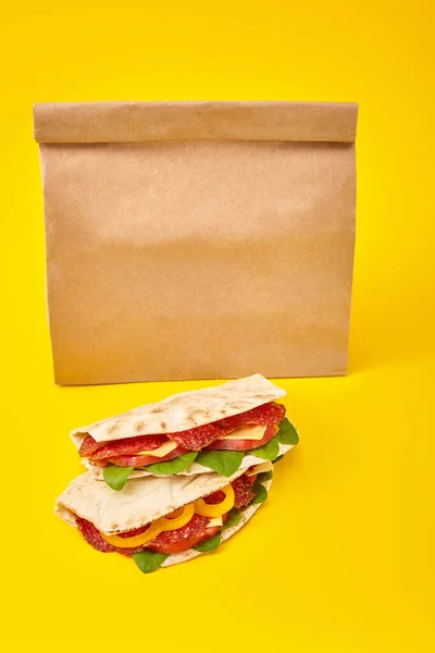 Fresh sandwiches with salami, pita, vegetables and cheese on yellow background near paper takeaway bag — Stock Photo