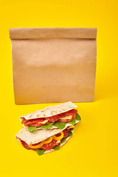 Bocadillos frescos con salami, pita, verduras y queso sobre fondo amarillo cerca de una bolsa de papel para llevar - foto de stock