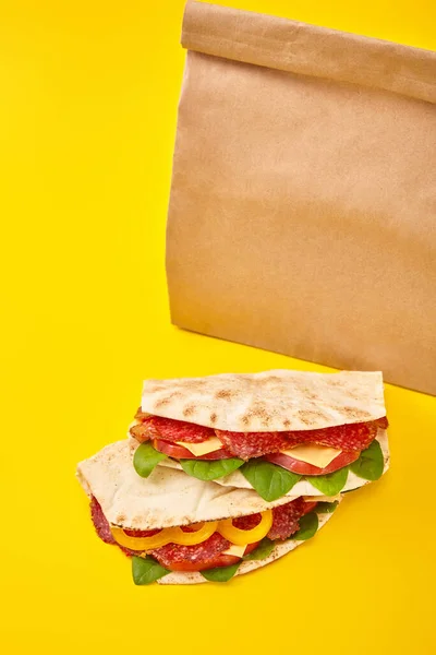 Bocadillos frescos con salami, pita, verduras y queso sobre fondo amarillo cerca de una bolsa de papel para llevar - foto de stock