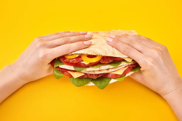 Cropped view of woman holding fresh sandwich with salami, pita, vegetables and cheese on yellow background — Stock Photo