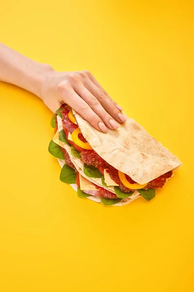Обрезанный вид женщины, держащей свежий сэндвич с салями, пита, овощи и сыр на желтом фоне — стоковое фото