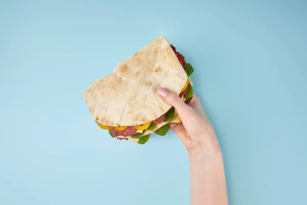Cropped view of woman holding fresh sandwich with salami, pita, vegetables and cheese on blue background — Stock Photo