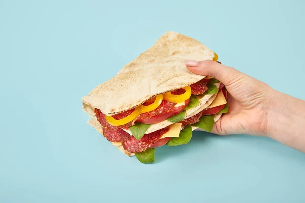 Vista recortada de la mujer sosteniendo sándwich fresco con salami, pita, verduras y queso sobre fondo azul - foto de stock