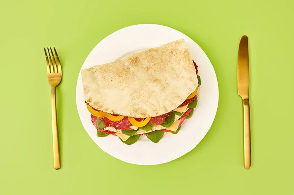 Vista superior de sándwich fresco con salami, pita, verduras y queso servido en el plato cerca de tenedor de oro y cuchillo sobre fondo verde - foto de stock