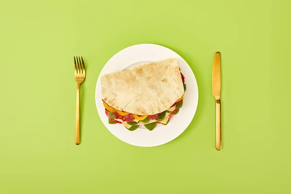 Top view of fresh sandwich with salami, pita, vegetables and cheese served on plate near golden fork and knife on green background — Stock Photo