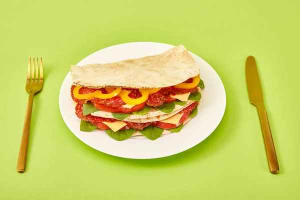 Fresh sandwich with salami, pita, vegetables and cheese served on plate near golden fork and knife on green background — Stock Photo