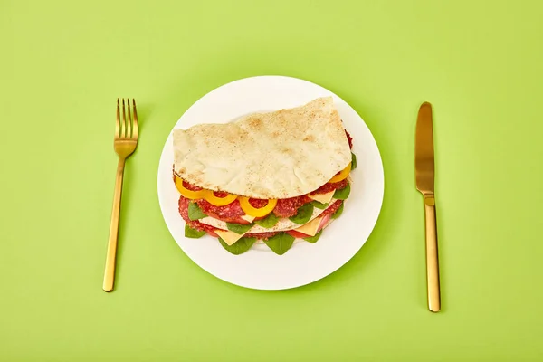 Fresh sandwich with salami, pita, vegetables and cheese served on plate near golden fork and knife on green background — Stock Photo