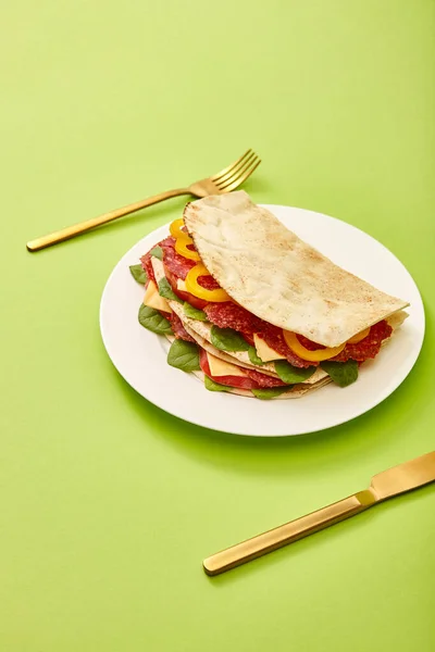 Fresh sandwich with salami, pita, vegetables and cheese served on plate near golden fork and knife on green background — Stock Photo