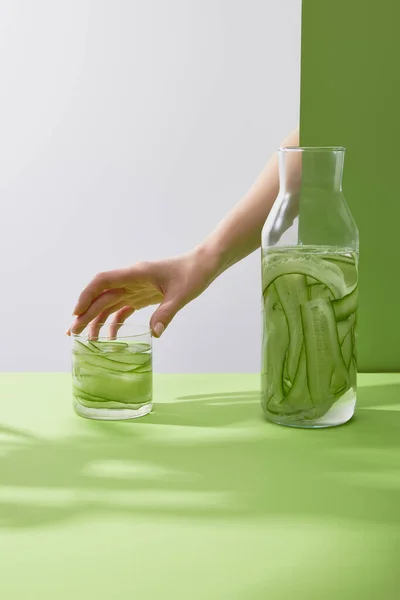 Vue recadrée de la main féminine touchant le verre avec une boisson faite de concombres tranchés sur fond gris et vert — Photo de stock