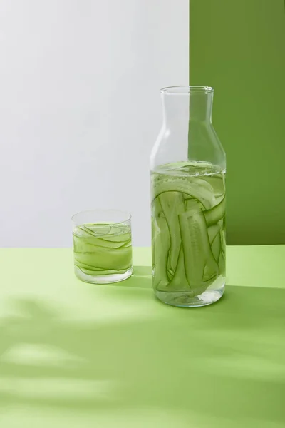 Bottle and glass with fresh drink made of sliced cucumbers on grey and green background — Stock Photo