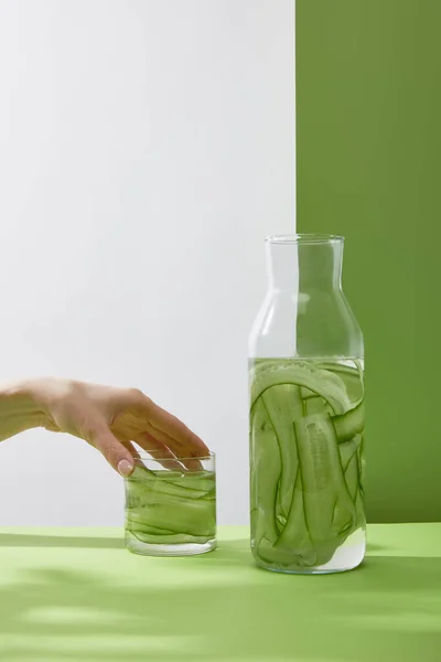 Vista recortada de la mano femenina tocando vidrio con bebida fresca hecha de pepinos en rodajas y botella sobre fondo gris y verde - foto de stock