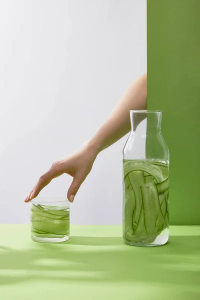 Vista recortada de la mano femenina tocando vidrio con bebida fresca hecha de pepinos en rodajas y botella sobre fondo gris y verde - foto de stock