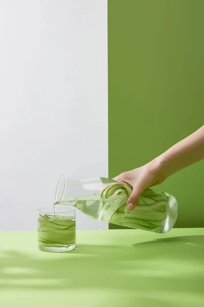 Vista recortada de la mano femenina vertiendo agua de la botella en vidrio con pepinos en rodajas sobre fondo gris y verde - foto de stock