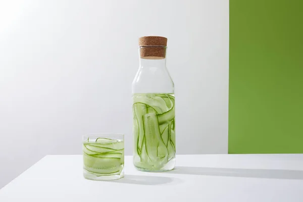 Bottle with cork and glass with drink made of sliced cucumbers on grey and green background — Stock Photo