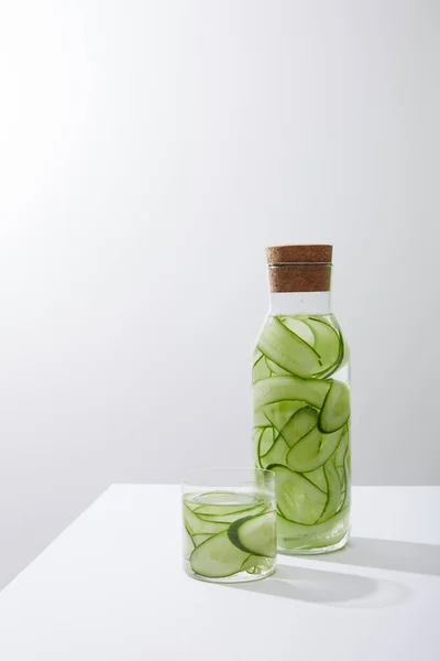 Bicchiere e bottiglia con tappo riempito con acqua e cetrioli a fette su fondo grigio — Foto stock