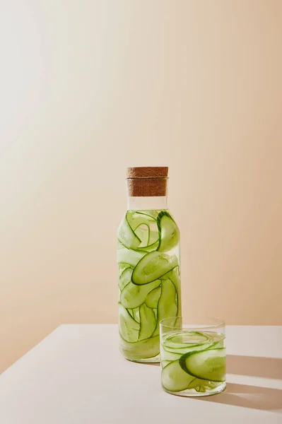 Vidrio y botella con corcho lleno de agua y pepinos en rodajas sobre la mesa sobre fondo beige - foto de stock