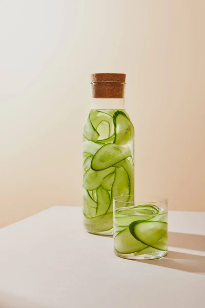 Glass and bottle with cork filled with fresh water and sliced cucumbers on beige background — Stock Photo