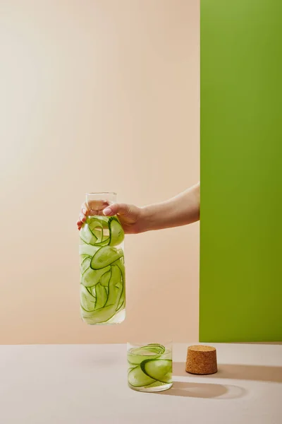 Cropped view of female hand holding bottle filled with water and sliced cucumbers on beige and green background — Stock Photo