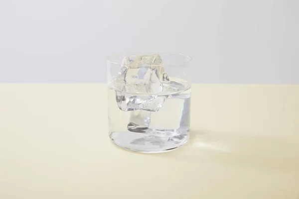 Glass with water and ice cubes on yellow surface isolated on grey — Stock Photo