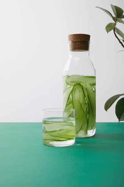 Bouteille avec du liège et du verre avec de l'eau et des concombres tranchés et des feuilles de plantes sur une surface verte isolée sur gris — Photo de stock