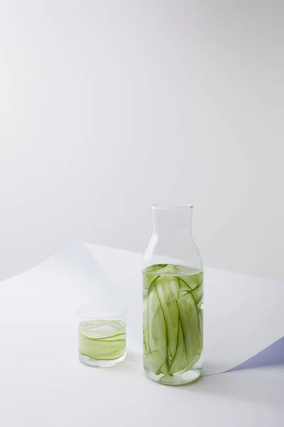Vista de alto ángulo de botella y vidrio con agua y pepinos en rodajas en la superficie de papel blanco aislado en gris - foto de stock