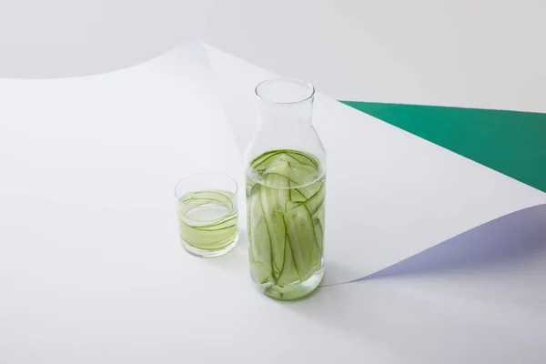 High angle view of bottle and glass with water and sliced cucumbers on white paper surface on grey and green background — Stock Photo