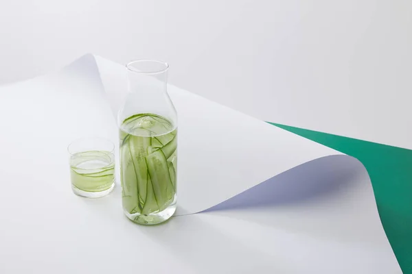 High angle view of bottle and glass with fresh drink and sliced cucumbers on white paper surface on grey and green background — Stock Photo