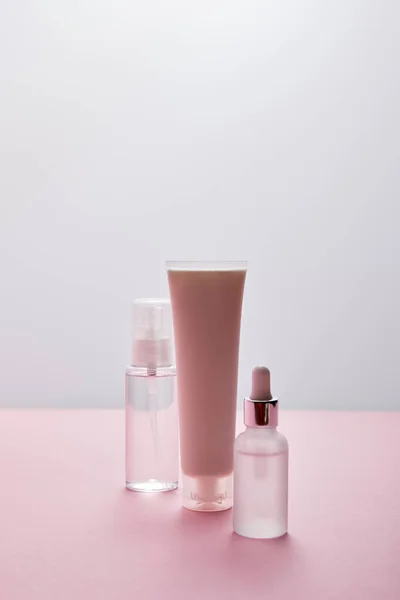 Set de cosméticos de tubo crema, spray con líquido y botella de suero sobre fondo rosa y gris - foto de stock