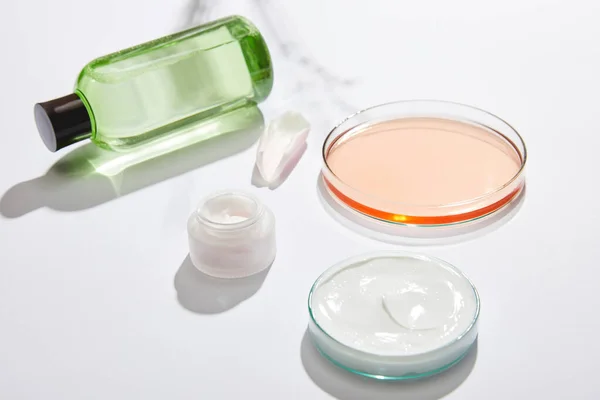 Bottiglia cosmetica con liquido verde, vaso con crema cosmetica bianca, bicchieri da laboratorio con liquido e petalo di rosa su fondo bianco con ombra — Foto stock