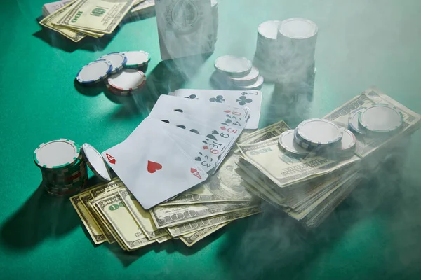 Vue en angle élevé des cartes à jouer, des billets en dollars et des jetons de casino avec de la fumée autour sur vert — Photo de stock