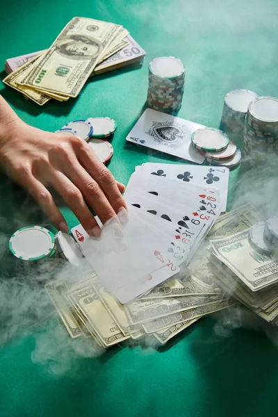 Vista recortada de la mano femenina con naipes, dinero y fichas de casino con humo alrededor en verde - foto de stock