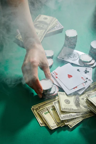 Cropped view of female hand with casino tokens, playing cards and money with smoke around on green background — Stock Photo