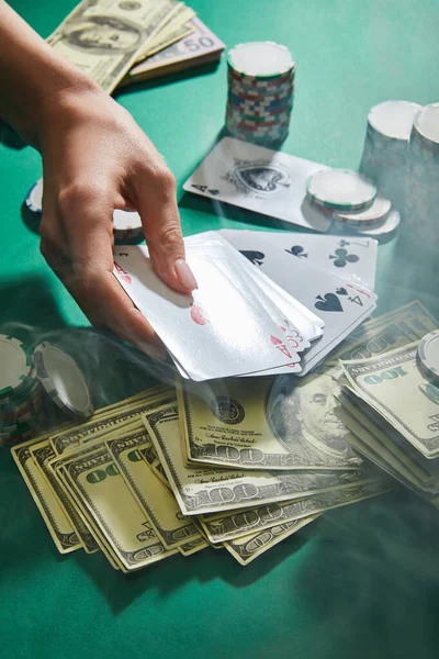Cropped view of woman holding playing cards near dollar banknotes and casino tokens with smoke around on green — Stock Photo