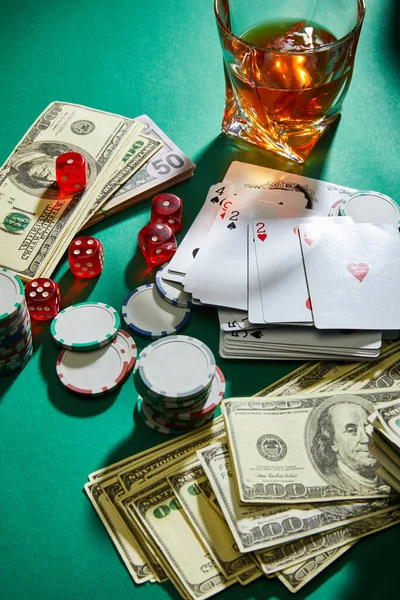 Vista de alto ángulo del dinero, jugando a las cartas, dados y fichas de casino cerca de un vaso de coñac en verde - foto de stock