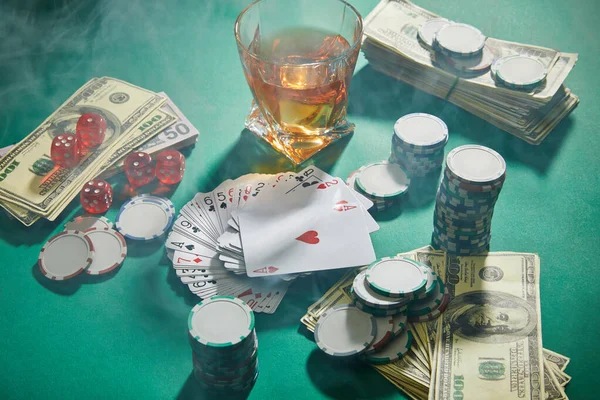 High angle view of money, playing cards, dice and casino tokens near glass of cognac with smoke around on green — Stock Photo