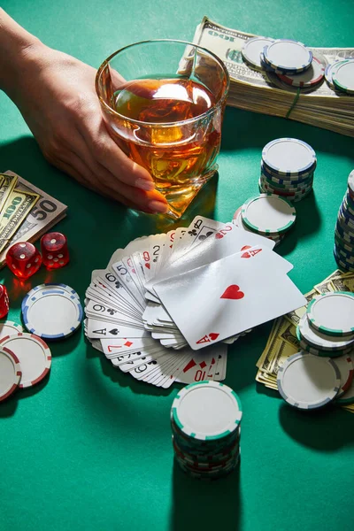 Vista recortada de la mujer sosteniendo un vaso de coñac cerca del dinero, jugando a las cartas, dados y fichas de casino en verde - foto de stock