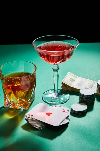Glasses of cognac and cocktail near dollar banknotes, casino tokens and playing cards on green surface isolated on black — Stock Photo