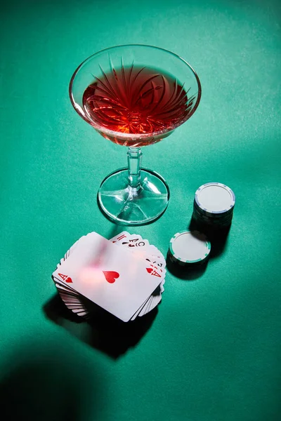 Vista de ángulo alto de vaso de cóctel cerca de jugar a las cartas y fichas de casino en verde - foto de stock