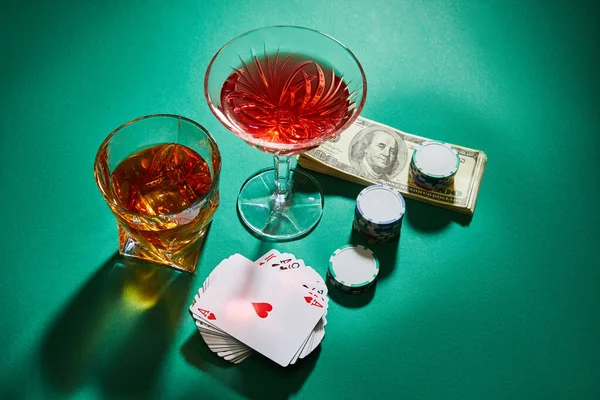 High angle view of glasses of cognac and cocktail near dollar banknotes, casino tokens and playing cards on green — Stock Photo