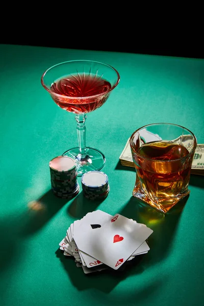 Vue grand angle des verres de cognac et cocktail près de l'argent, des jetons de casino et des cartes à jouer sur une surface verte isolée sur du noir — Photo de stock