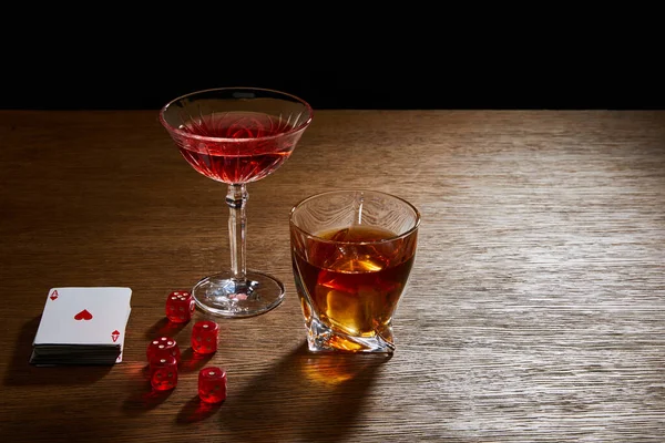 Glasses of cocktail and cognac near deck of cards and dice on wooden surface isolated on black — Stock Photo