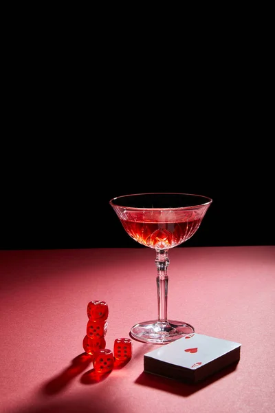 Glass of cocktail near deck of cards and dice on red surface isolated on black — Stock Photo