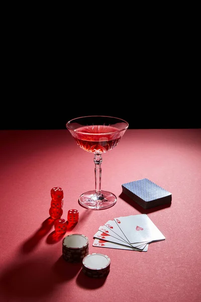 Glass of cocktail near playing cards, casino chips and dice on red surface isolated on black — Stock Photo