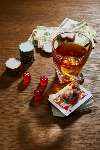 Vue en angle élevé du verre de cognac près des billets en dollars, des cartes à jouer, des jetons de casino et des dés sur fond en bois — Photo de stock